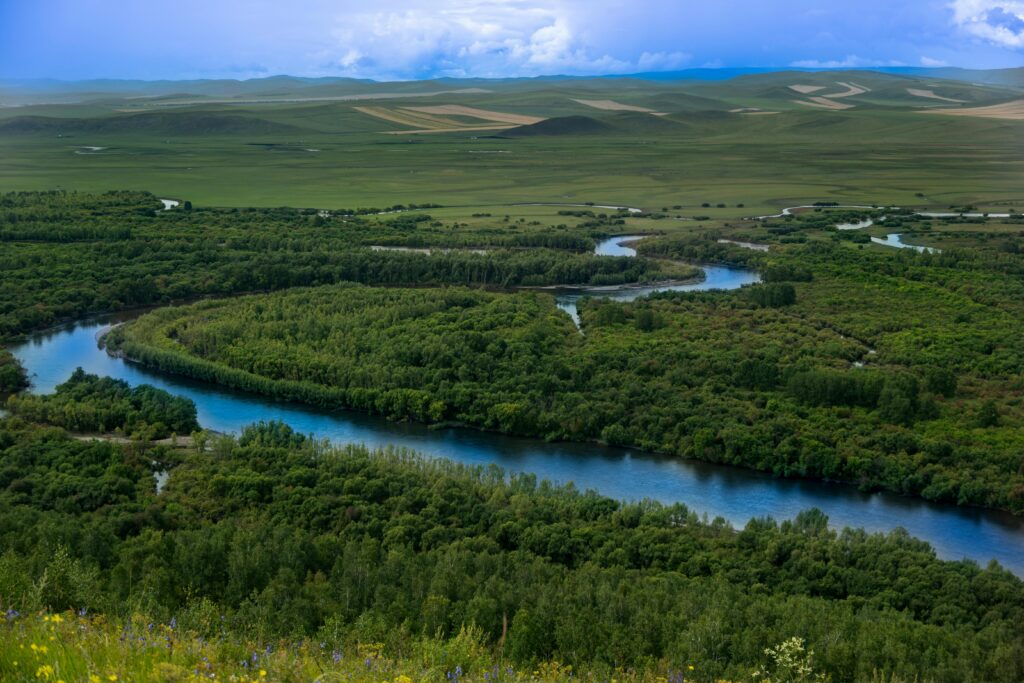 Aerial view of a winding river flowing through lush green forests and fields under a bright sky.